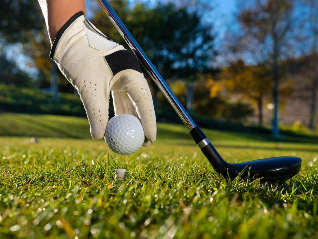 Golf glove wearing hand placing ball on tee with club on sunny day.